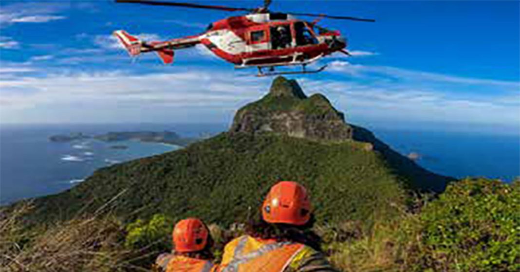 Red and white helicopter hovering in the sky with a mountain in the distance and three people in orange safety gear in the foreground